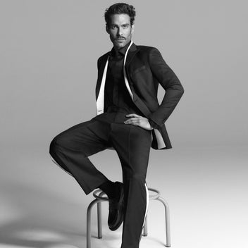 Man in a black suit sitting on a stool against a gray background