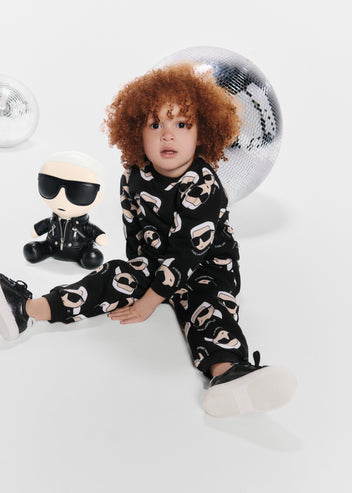 Child wearing a black outfit with white patterns, sitting on a white surface with a toy and disco ball in the background.